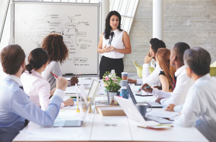 Woman giving presentation on whiteboard in boardroom meeting to seven office worker colleagues